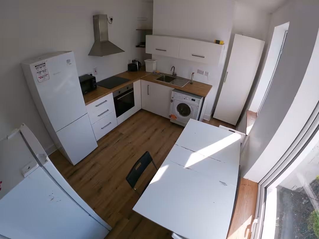 Compact kitchen with white cabinets, appliances, table, and chair. Bright natural light.