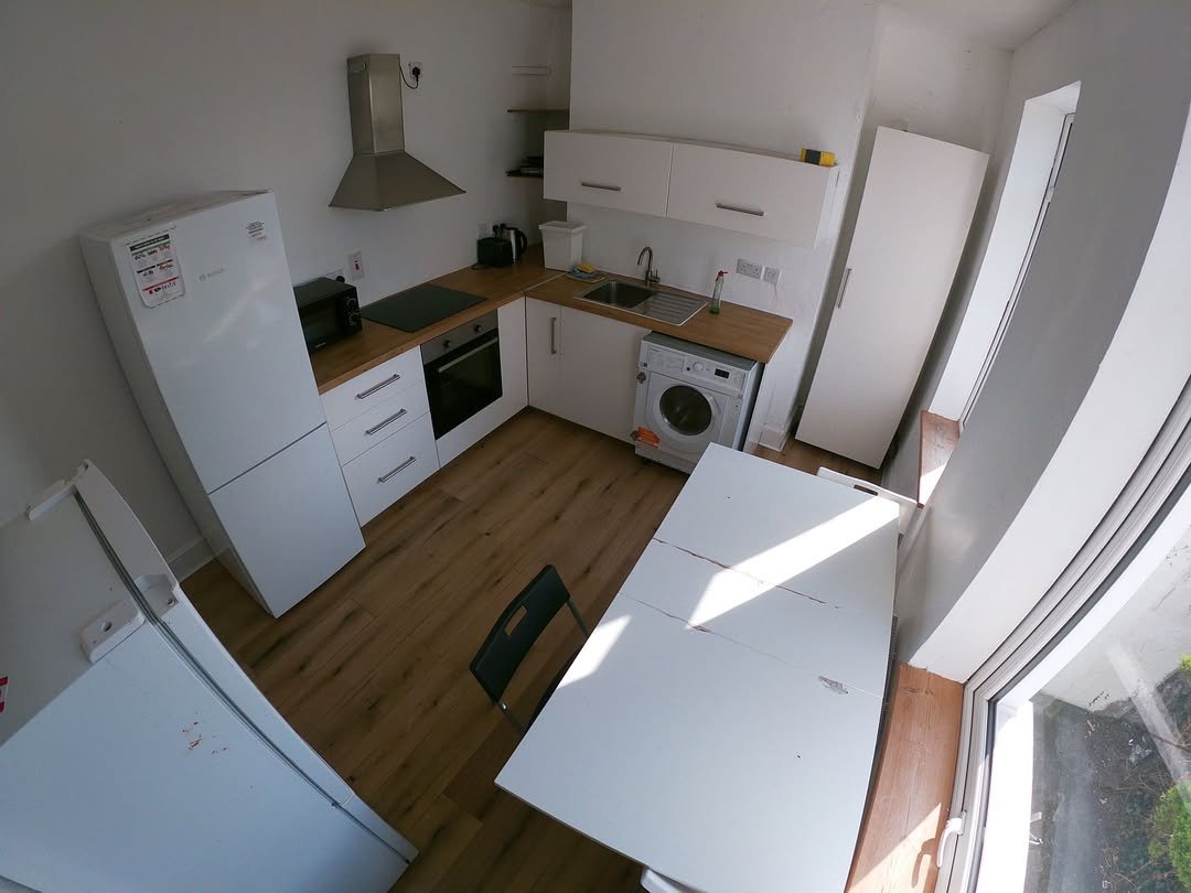 Compact kitchen with white cabinets, appliances, table, and chair. Bright natural light.