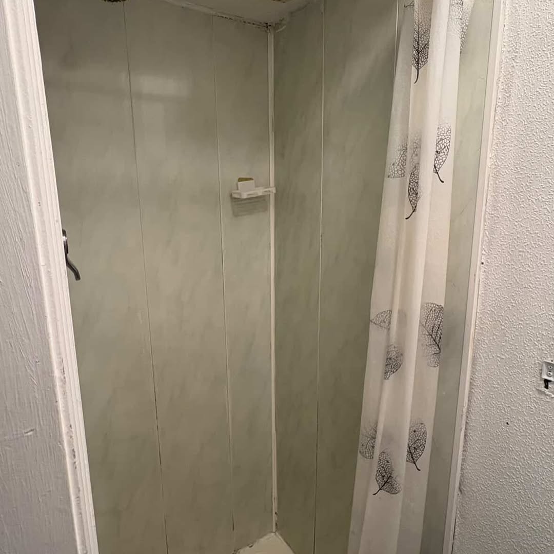 Small shower stall with white paneling and leaf-patterned curtain.