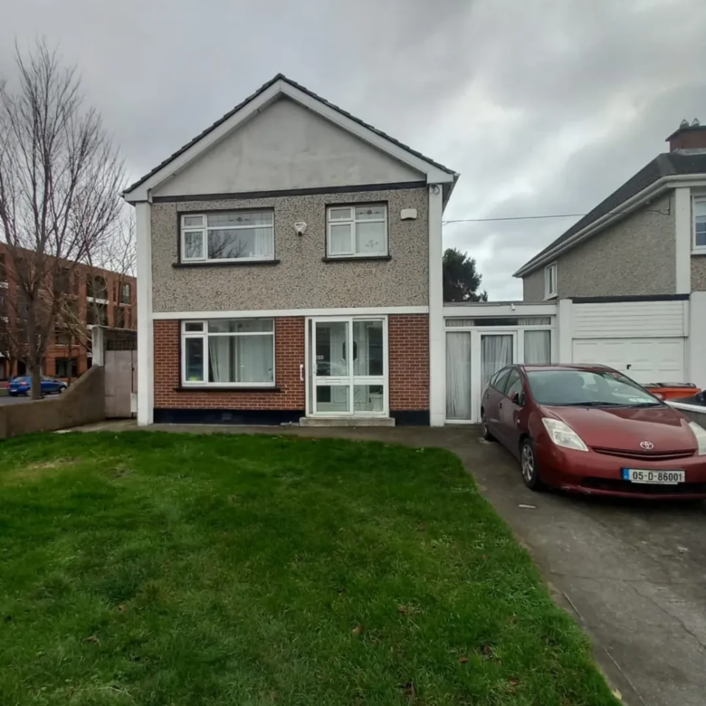 Two-story house with brick accents, green lawn, and a red car parked in the driveway under a cloudy sky.