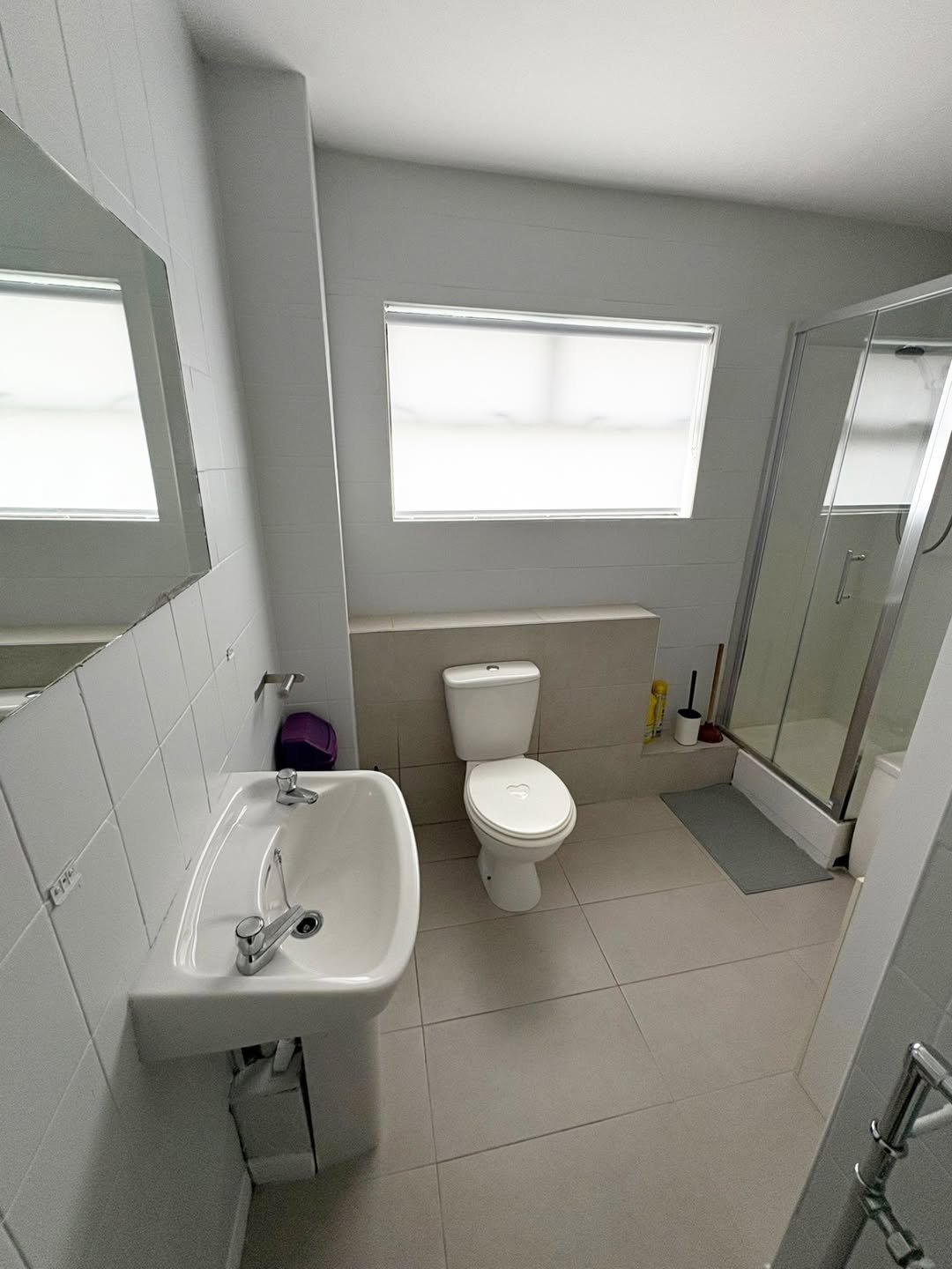 Bathroom with white toilet, sink, shower, and neutral tile flooring.