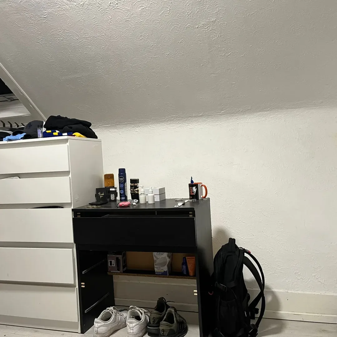 Bedroom dresser and desk with toiletries, shoes, and backpack against a white wall.