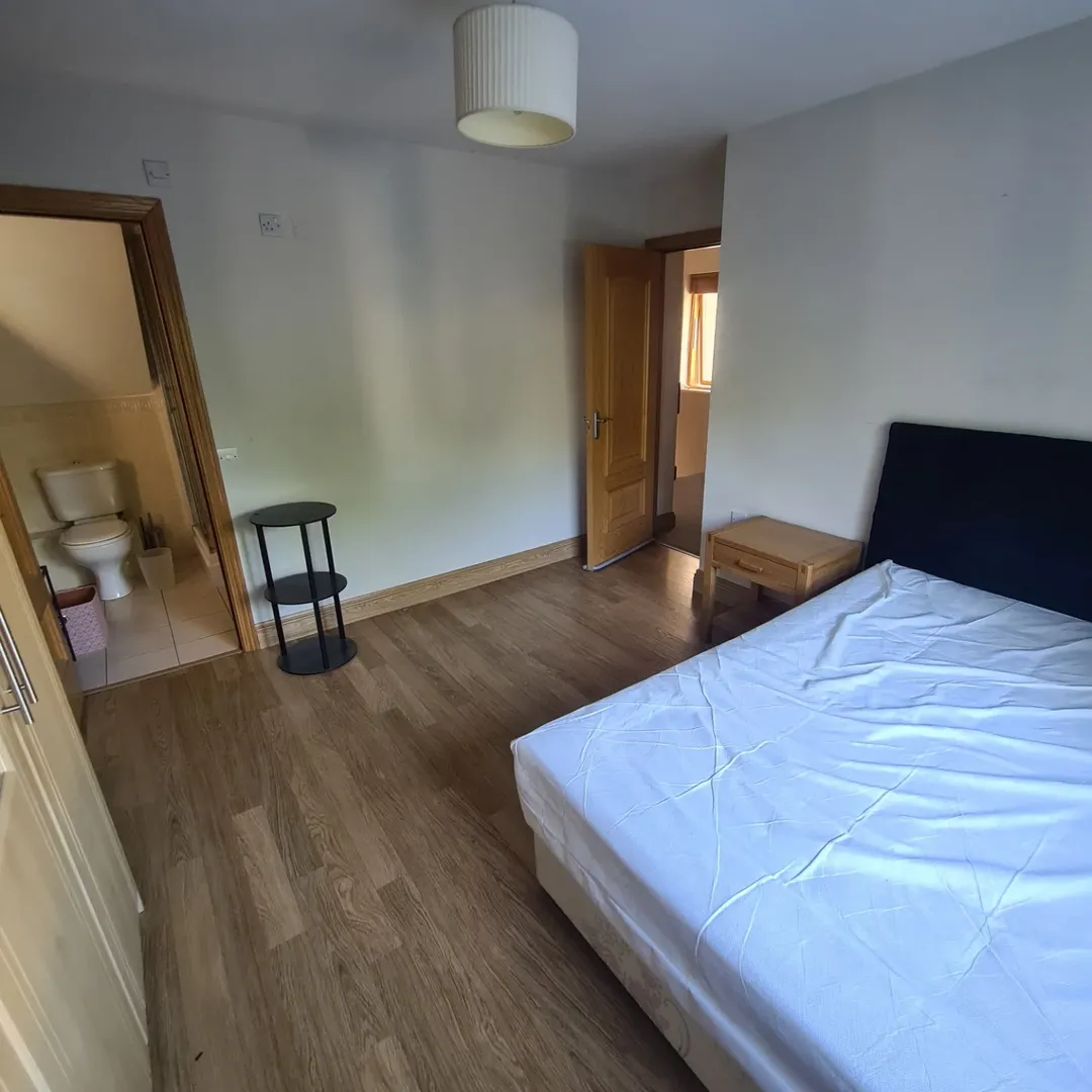 Bedroom with bed, ensuite bathroom, and small side table. Neutral color scheme.