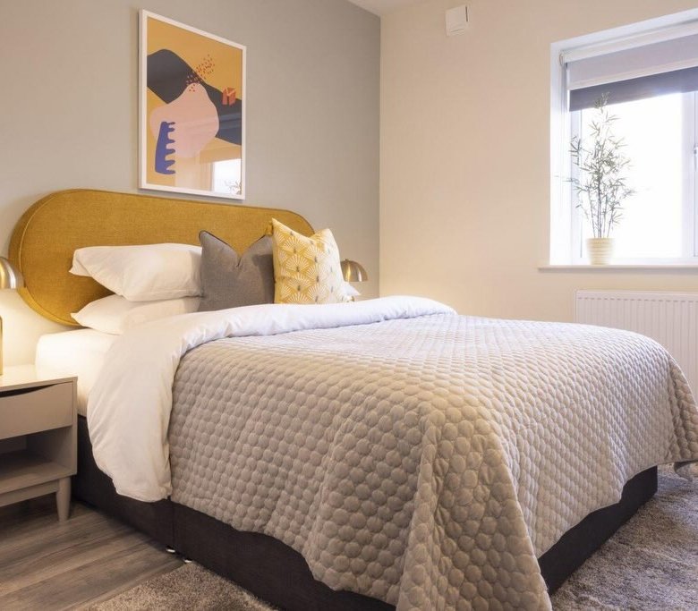 Modern bedroom with yellow headboard, grey quilt, nightstand, and abstract art on the wall.