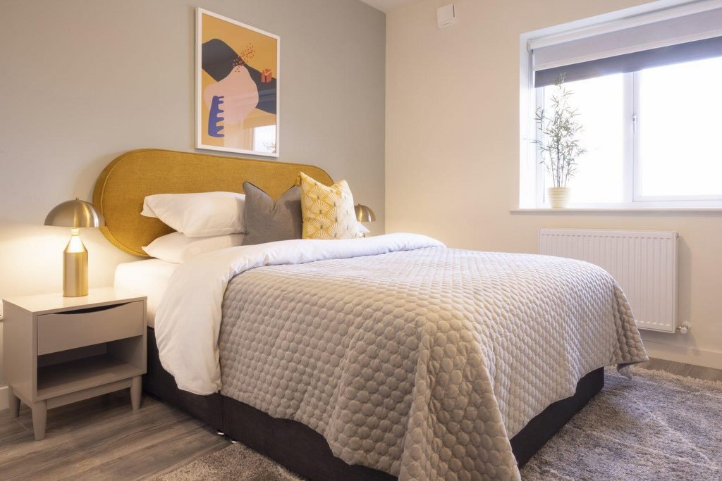 Modern bedroom with yellow headboard, grey quilt, nightstand, and abstract art on the wall.