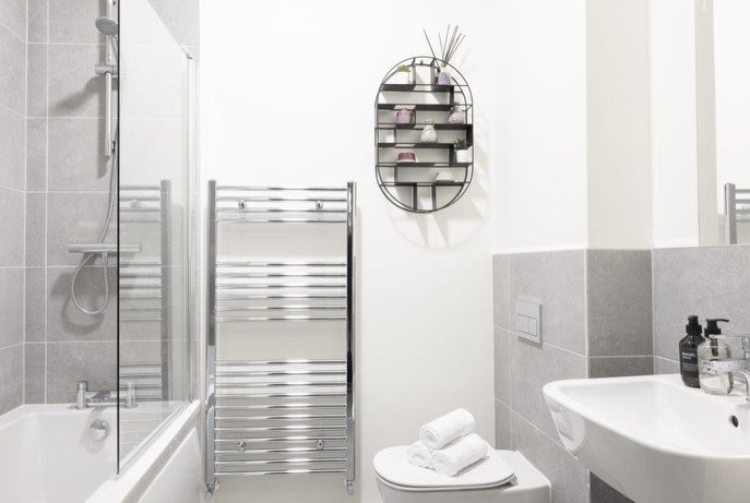 Modern bathroom with gray tile, chrome towel warmer, oval shelf, toilet, and sink. White walls and rolled towels.