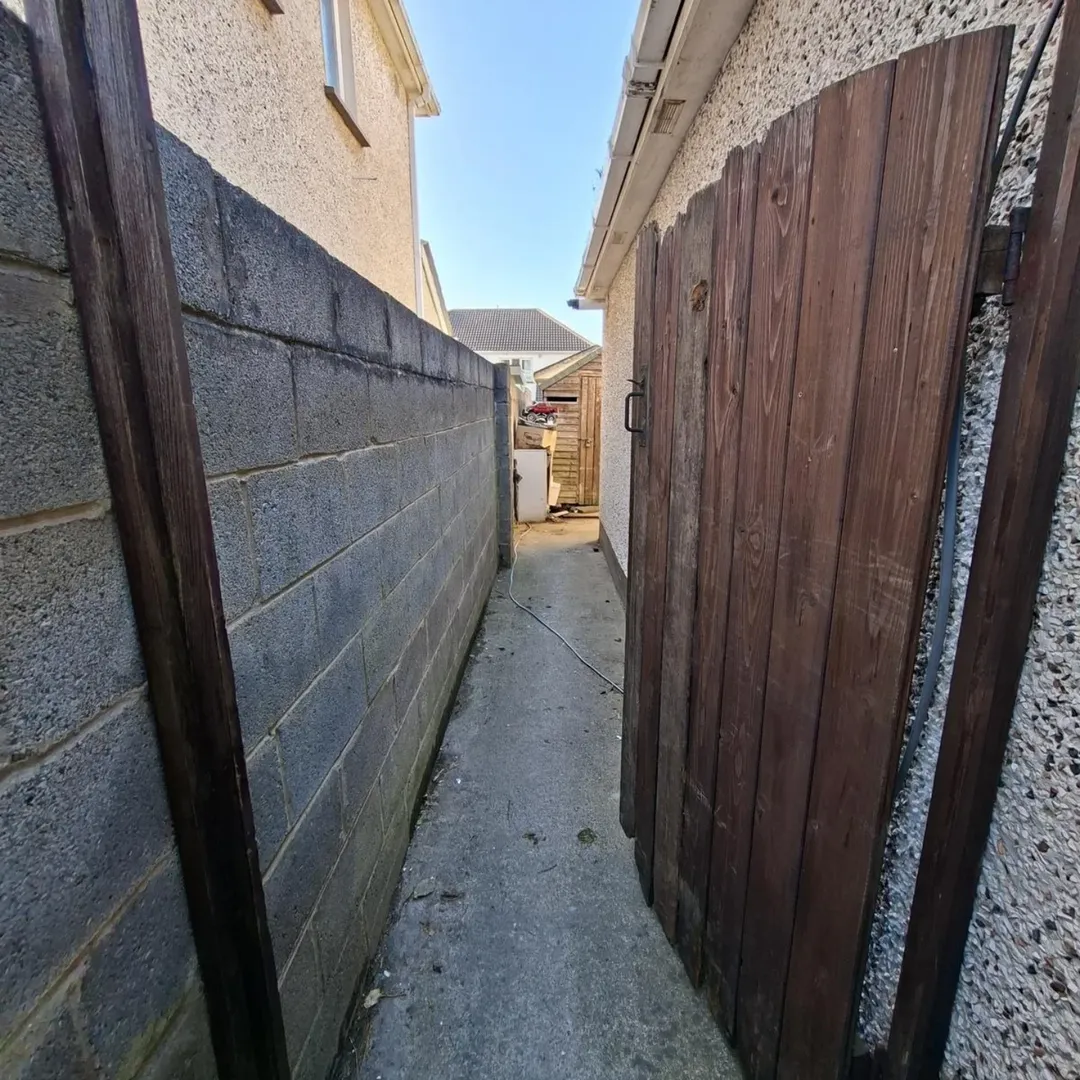 Narrow alleyway between house wall and wooden gate leading to backyard shed.