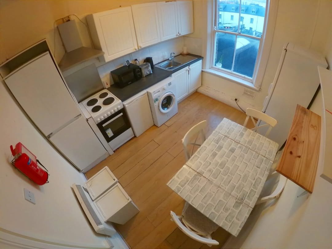 Compact kitchen with white cabinets, appliances, dining table, and chairs in a small apartment setting.