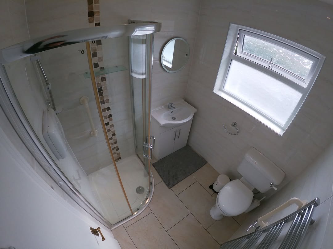 Small bathroom with corner shower, toilet, sink, round mirror, and a window. Neutral tile and fixtures.