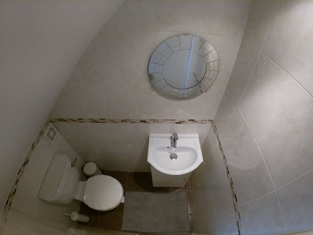 Small bathroom with toilet, sink, round mirror, and tiled walls under a sloped ceiling.