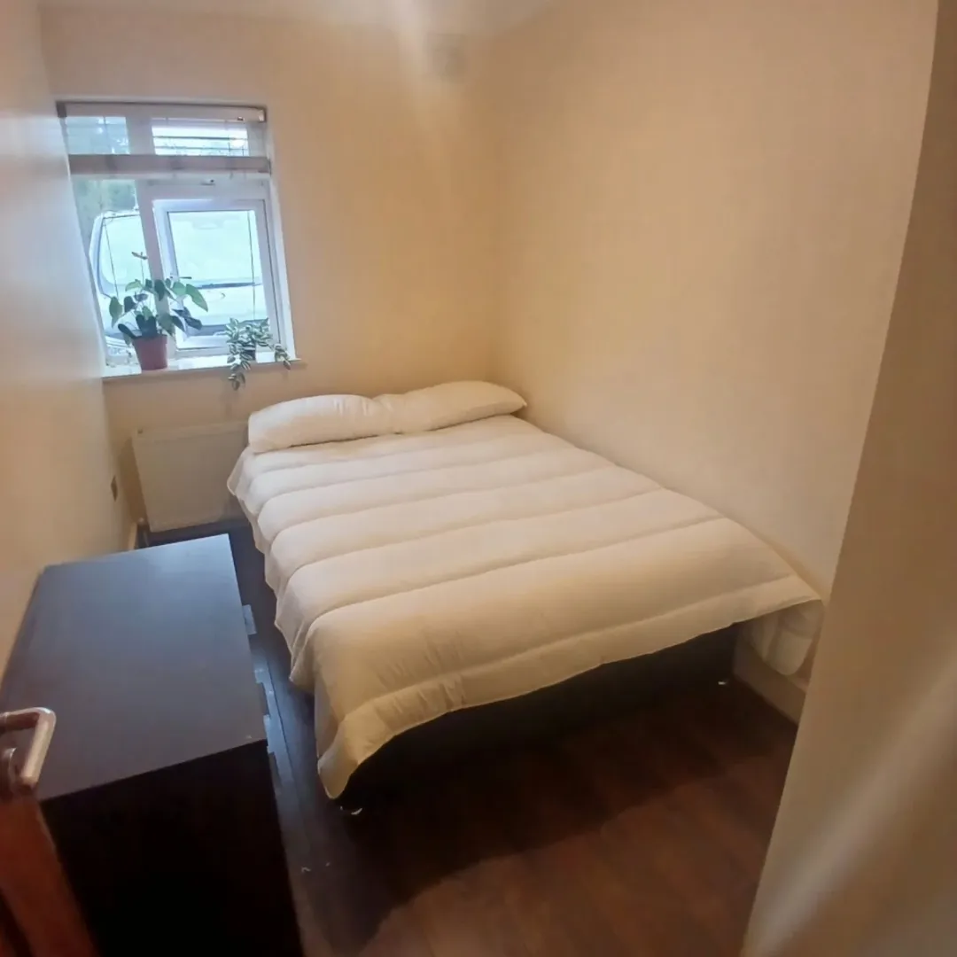 Small bedroom with double bed, dark dresser, and window with plants.