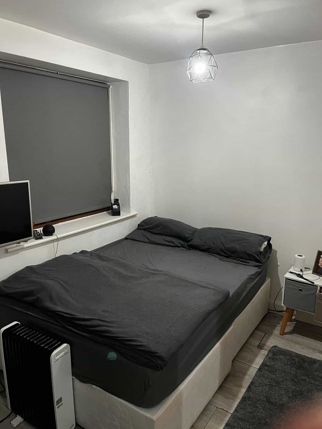 Small bedroom with gray bedding, window, bedside table, and heater. Minimalist interior design.