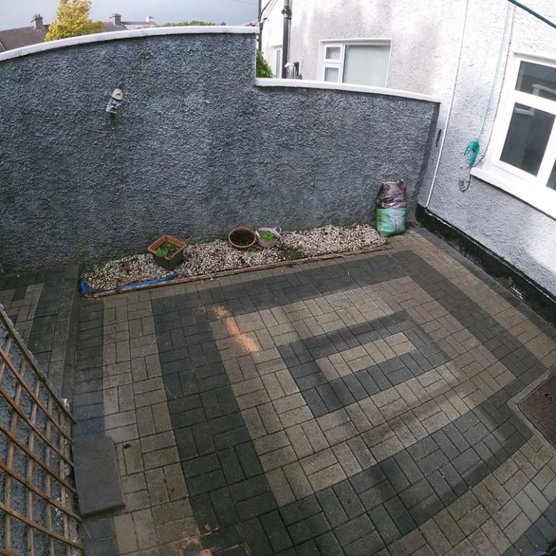 Small paved backyard patio with gray textured wall, potted plants, and a trellis fence.
