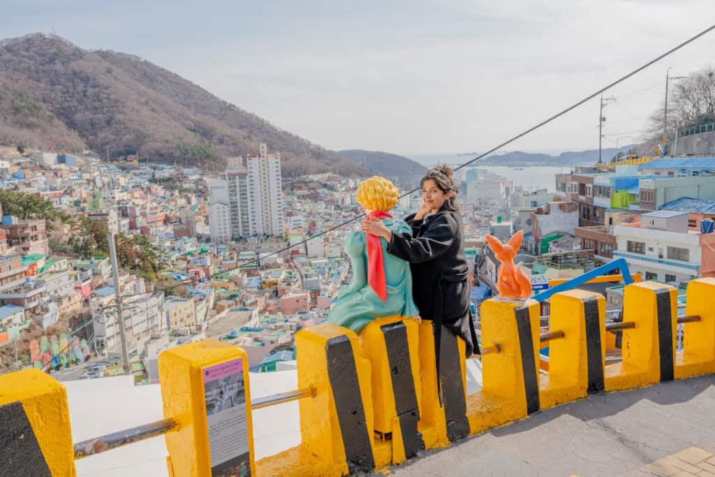 도시와 자연이 조화를 이루는 한국 촬영지, 인기 테마 포토존에서 사진 찍는 여성과 만화 인형, 드라이브와 여행을 즐기는 모습, 부산 또는 서울의 산과 바다 전망 포함.