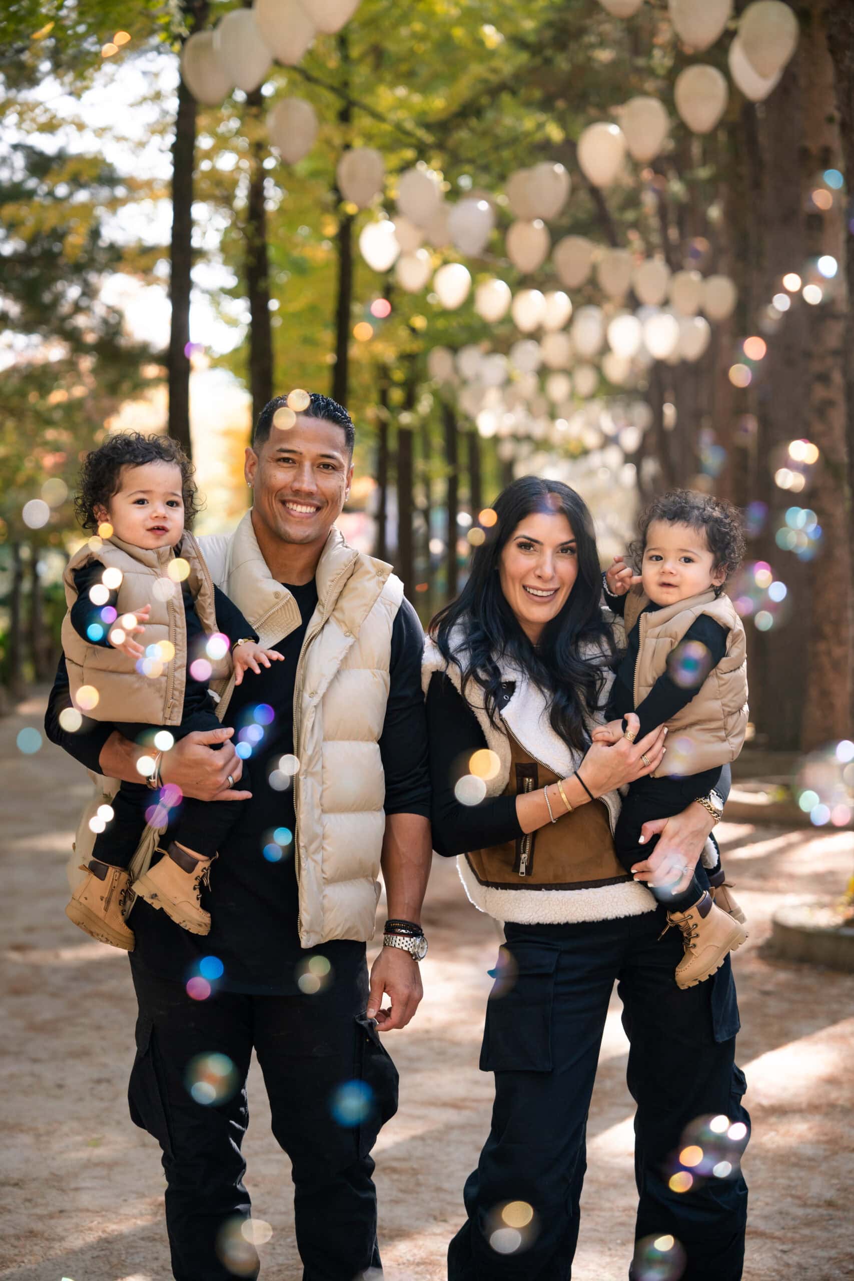 Family portrait in Korea during golden hour, capturing parents with young children in a scenic outdoor park with lanterns, vibrant autumn leaves, and bokeh lighting effects.