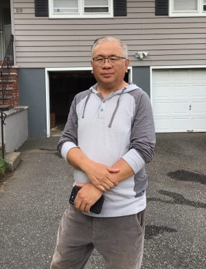 %Focuskw% | Good Tidings Plumbing, Heating &Amp; Cooling A man wearing glasses and a gray hoodie stands on a driveway in front of a house with a garage, holding a smartphone.