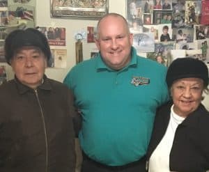 %Focuskw% | Good Tidings Plumbing, Heating &Amp; Cooling Three people stand close together indoors, posing and smiling for a photo in front of a wall covered with various photographs.