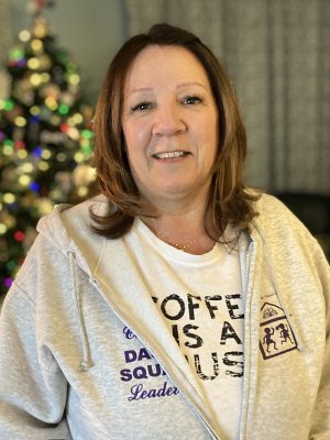 %Focuskw% | Good Tidings Plumbing, Heating &Amp; Cooling Woman with shoulder-length brown hair wearing a light hoodie and a printed T-shirt, standing indoors with a blurred Christmas tree in the background.