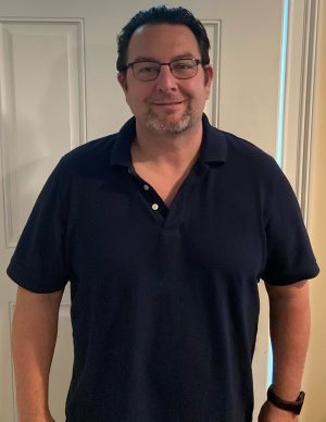 %Focuskw% | Good Tidings Plumbing, Heating &Amp; Cooling A man with glasses and short dark hair stands indoors, wearing a navy blue polo shirt, in front of a white door.
