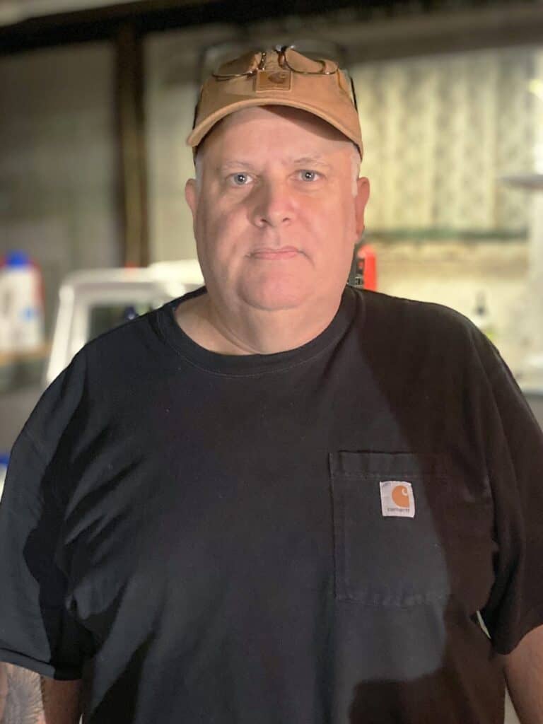 %Focuskw% | Good Tidings Plumbing, Heating &Amp; Cooling A man wearing a tan cap, glasses, and a black Carhartt t-shirt stands indoors in a dimly lit room with blurred background objects.