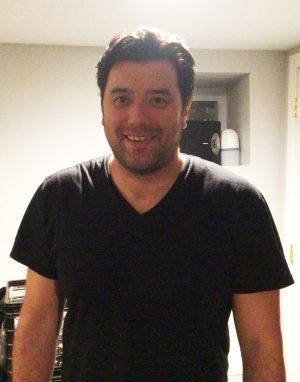 %Focuskw% | Good Tidings Plumbing, Heating &Amp; Cooling A man with dark hair and a short beard is standing indoors, smiling at the camera. He is wearing a black V-neck T-shirt, and the background features a shelf and a closed door.