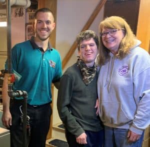 %Focuskw% | Good Tidings Plumbing, Heating &Amp; Cooling Three people stand indoors near a staircase; one man in a green shirt, another man in a gray sweater, and a woman in a gray hoodie, all smiling at the camera.