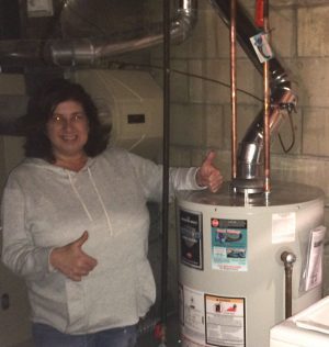 %Focuskw% | Good Tidings Plumbing, Heating &Amp; Cooling A woman stands next to a water heater in a basement, giving a thumbs-up gesture and smiling at the camera.