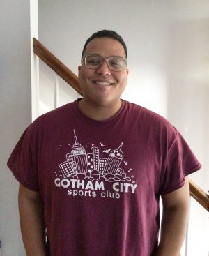 %Focuskw% | Good Tidings Plumbing, Heating &Amp; Cooling A person wearing glasses and a maroon "Gotham City Sports Club" t-shirt stands indoors in front of a staircase, smiling at the camera.