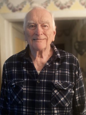 %Focuskw% | Good Tidings Plumbing, Heating &Amp; Cooling Elderly man with gray hair wearing a blue and white plaid shirt stands indoors, smiling slightly at the camera.