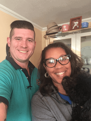 %Focuskw% | Good Tidings Plumbing, Heating &Amp; Cooling A man and woman smile at the camera indoors; the woman holds a small black dog. Shelves with cups and a framed photo are in the background.