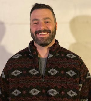 %Focuskw% | Good Tidings Plumbing, Heating &Amp; Cooling A man with short hair and a beard is smiling, wearing a patterned fleece jacket, standing in front of a plain light-colored wall.