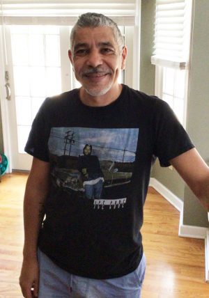 %Focuskw% | Good Tidings Plumbing, Heating &Amp; Cooling Middle-aged man with short gray hair and a beard wearing a black graphic t-shirt, standing indoors by a window with blinds and a glass door in the background.