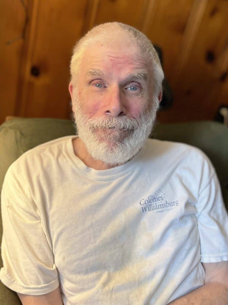 %Focuskw% | Good Tidings Plumbing, Heating &Amp; Cooling An older man with a white beard and short hair sits on a couch, wearing a white "Colonies: Williamsburg" t-shirt. Wood panel wall in the background.