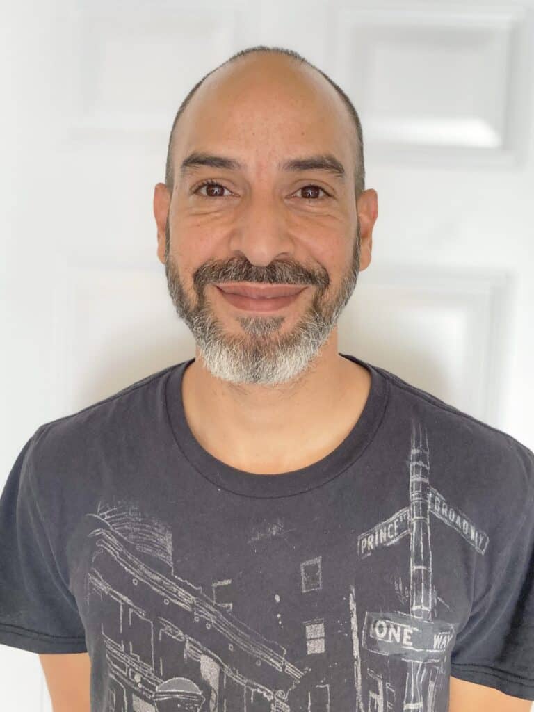 %Focuskw% | Good Tidings Plumbing, Heating &Amp; Cooling A man with a shaved head and short beard smiles at the camera, wearing a black graphic t-shirt, standing in front of a white door.