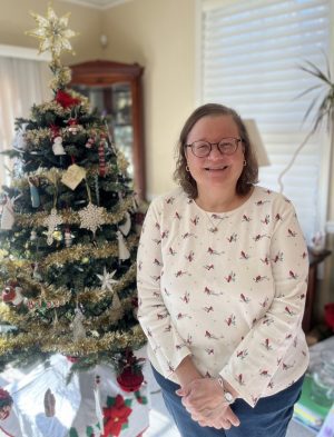 %Focuskw% | Good Tidings Plumbing, Heating &Amp; Cooling A woman wearing glasses and a white patterned sweater stands smiling beside a decorated Christmas tree in a well-lit room.