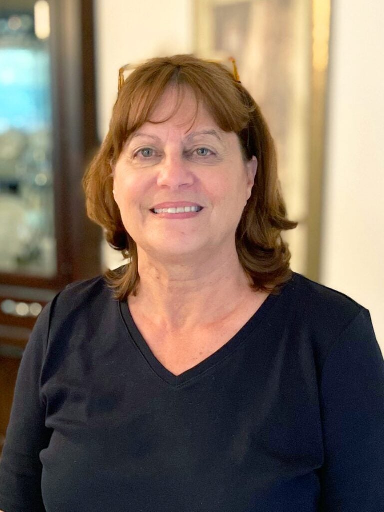 %Focuskw% | Good Tidings Plumbing, Heating &Amp; Cooling A woman with shoulder-length brown hair and glasses on her head is smiling, wearing a black v-neck shirt, standing indoors with a blurred background.