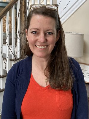 %Focuskw% | Good Tidings Plumbing, Heating &Amp; Cooling A woman with long brown hair wearing an orange top and navy cardigan stands indoors, smiling at the camera. A staircase and lamp are visible in the background.