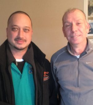%Focuskw% | Good Tidings Plumbing, Heating &Amp; Cooling Two men standing side by side indoors, both facing the camera and smiling slightly. One wears a teal shirt with a black jacket, the other wears a light gray zip-up sweater.