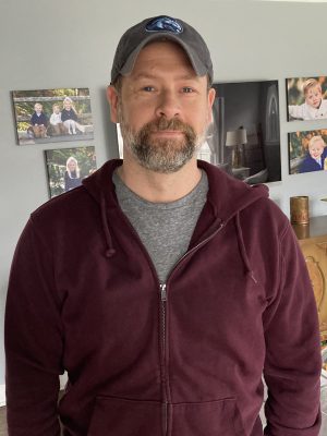 %Focuskw% | Good Tidings Plumbing, Heating &Amp; Cooling A man with a beard wearing a gray cap, maroon zip-up hoodie, and gray t-shirt stands indoors with family photos on the wall behind him.