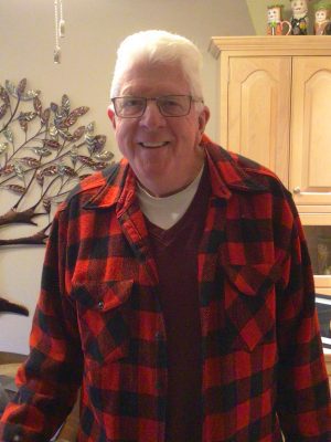 %Focuskw% | Good Tidings Plumbing, Heating &Amp; Cooling An older man with short white hair and glasses is smiling, wearing a red and black plaid shirt over a maroon sweater, standing indoors near a wooden cabinet and wall art.
