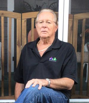 %Focuskw% | Good Tidings Plumbing, Heating &Amp; Cooling An older man with glasses and a necklace sits on a chair, wearing a black polo shirt and jeans, with a wooden fence and glass doors in the background.