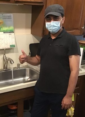 %Focuskw% | Good Tidings Plumbing, Heating &Amp; Cooling A man wearing a mask and cap stands by a kitchen sink with running water, giving a thumbs-up gesture.