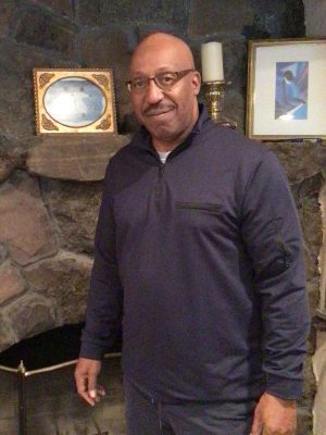 %Focuskw% | Good Tidings Plumbing, Heating &Amp; Cooling A man wearing glasses and a navy pullover stands in front of a stone fireplace with framed pictures and a candle on the mantel.