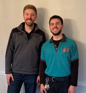 %Focuskw% | Good Tidings Plumbing, Heating &Amp; Cooling Two men standing indoors, smiling at the camera. One wears a gray quarter-zip pullover and jeans, the other wears a teal collared shirt with keys hanging from his belt.