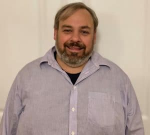 %Focuskw% | Good Tidings Plumbing, Heating &Amp; Cooling A man with gray hair and a beard, wearing a light purple button-up shirt, stands in front of a plain white background, smiling at the camera.