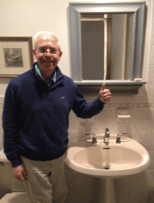 %Focuskw% | Good Tidings Plumbing, Heating &Amp; Cooling Older man gives a thumbs up while standing next to a bathroom sink and mirror, with a towel hanging on the wall and framed art in the background.