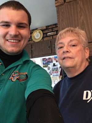 %Focuskw% | Good Tidings Plumbing, Heating &Amp; Cooling Two people, one younger and one older, stand indoors and smile at the camera. The younger person wears a green polo shirt; the older person wears a navy blue shirt.