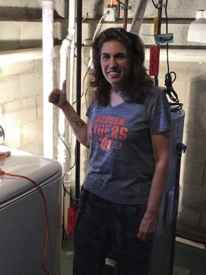 %Focuskw% | Good Tidings Plumbing, Heating &Amp; Cooling Woman stands in a basement next to a washing machine, giving a thumbs up. She is wearing an Auburn Tigers t-shirt and dark pants. Pipes and a water heater are visible in the background.