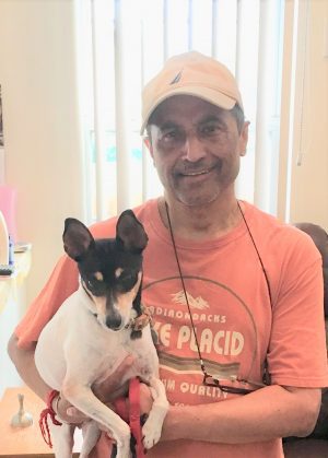 %Focuskw% | Good Tidings Plumbing, Heating &Amp; Cooling A man wearing a tan cap and orange t-shirt holds a small black and white dog with pointed ears indoors.