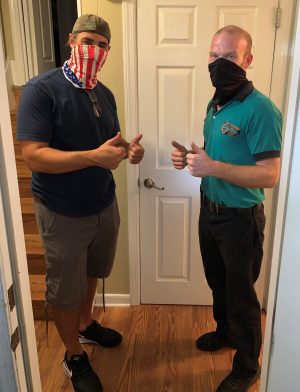 %Focuskw% | Good Tidings Plumbing, Heating &Amp; Cooling Two men wearing face coverings stand indoors, giving thumbs up. One wears a flag mask and cap; the other wears a black mask and teal shirt. They stand in front of a closed white door.